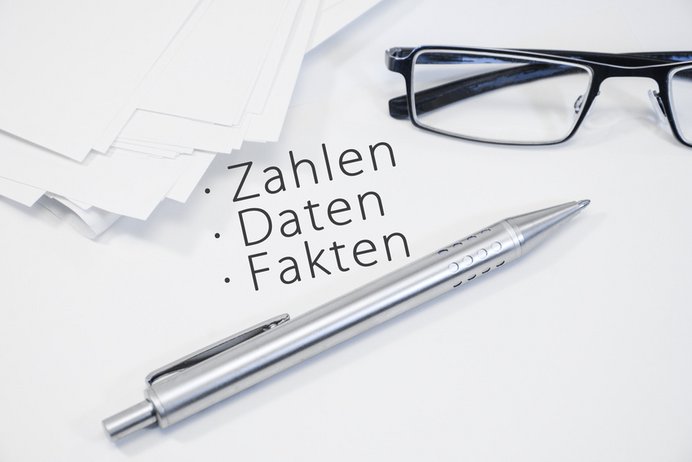 Brille, Kugelschreiber und mehrere weiße Blätter liegen auf einem Blatt Papier, auf dem "Zahlen, Daten, Fakten" steht.